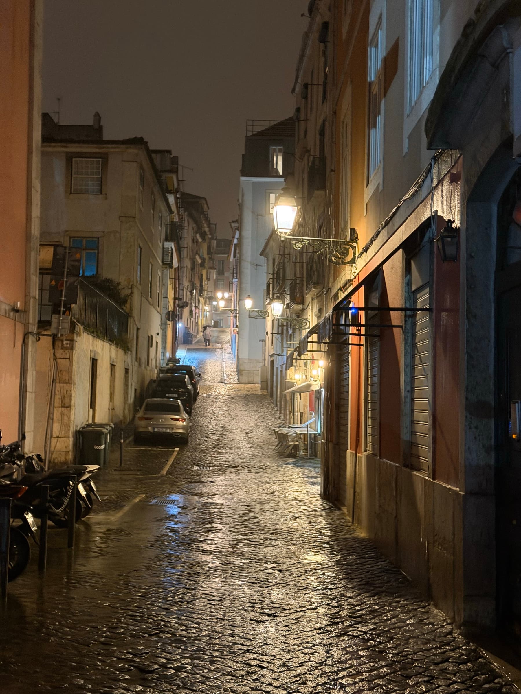 A wet Lisbon cobblestone street at night, lit by warm lamps and reflected light.