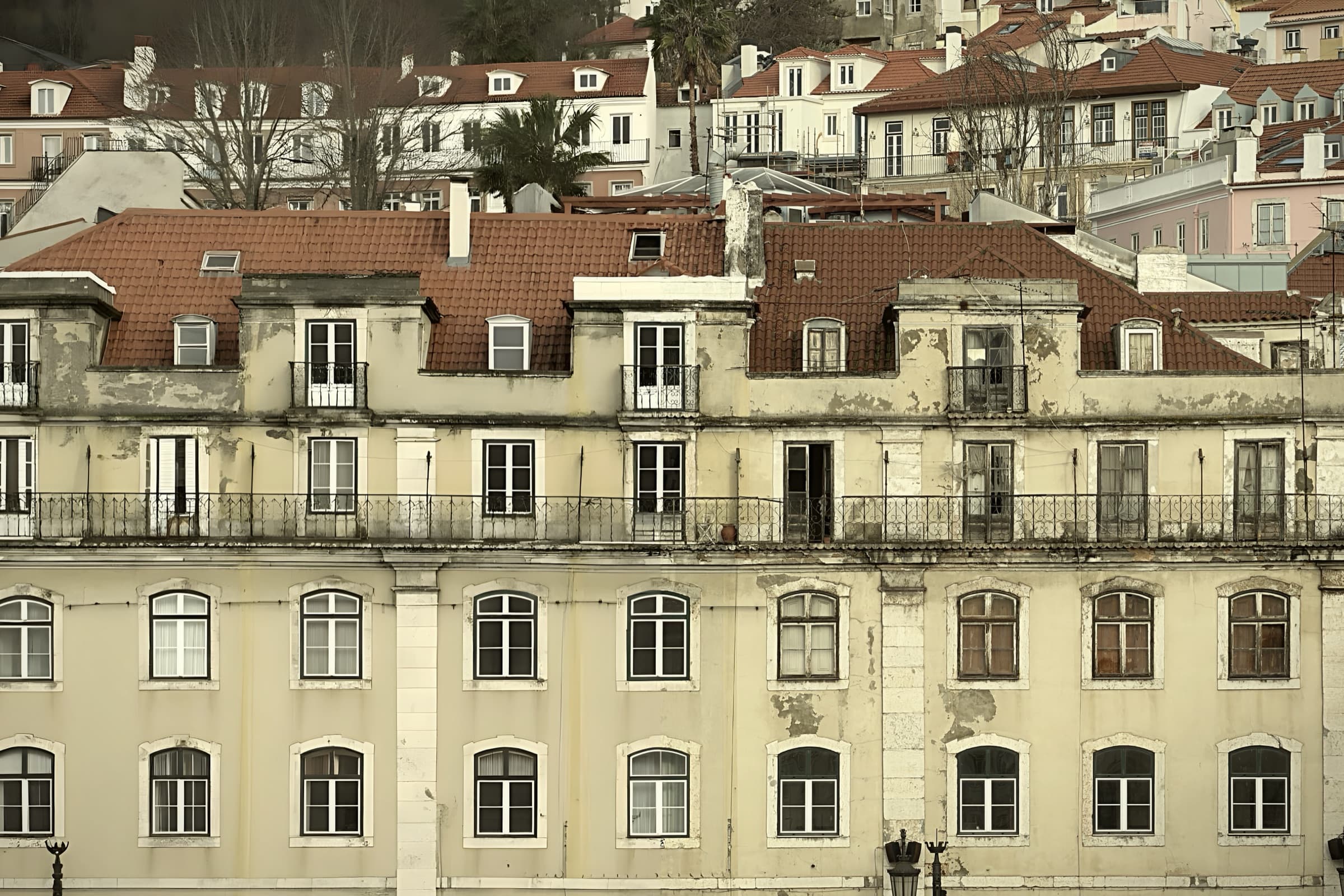 A layered Lisbon facade with worn cream walls, tiled roofs, balconies, and many windows.
